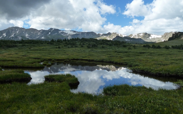 a rocky and snowy mountain ridge is reflected in the still waters of a pond